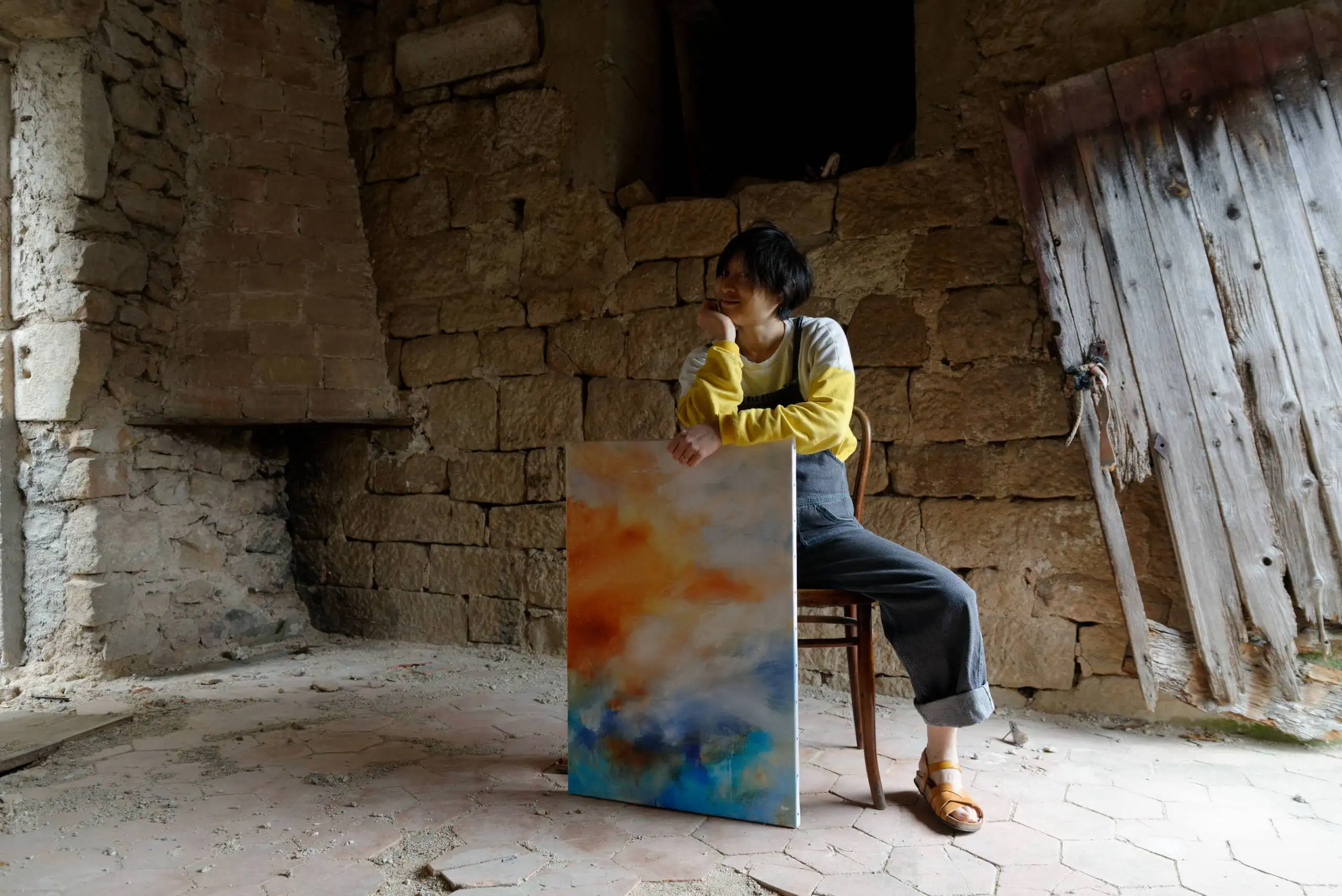 Portrait de l'artiste peintre Mai Khanh Pham To dans son atelier en Ardèche. Elle est assise sur une chaise devant une toile peinte à l'huile appelée l'Archipel, dans des tons orange et bleu azur.