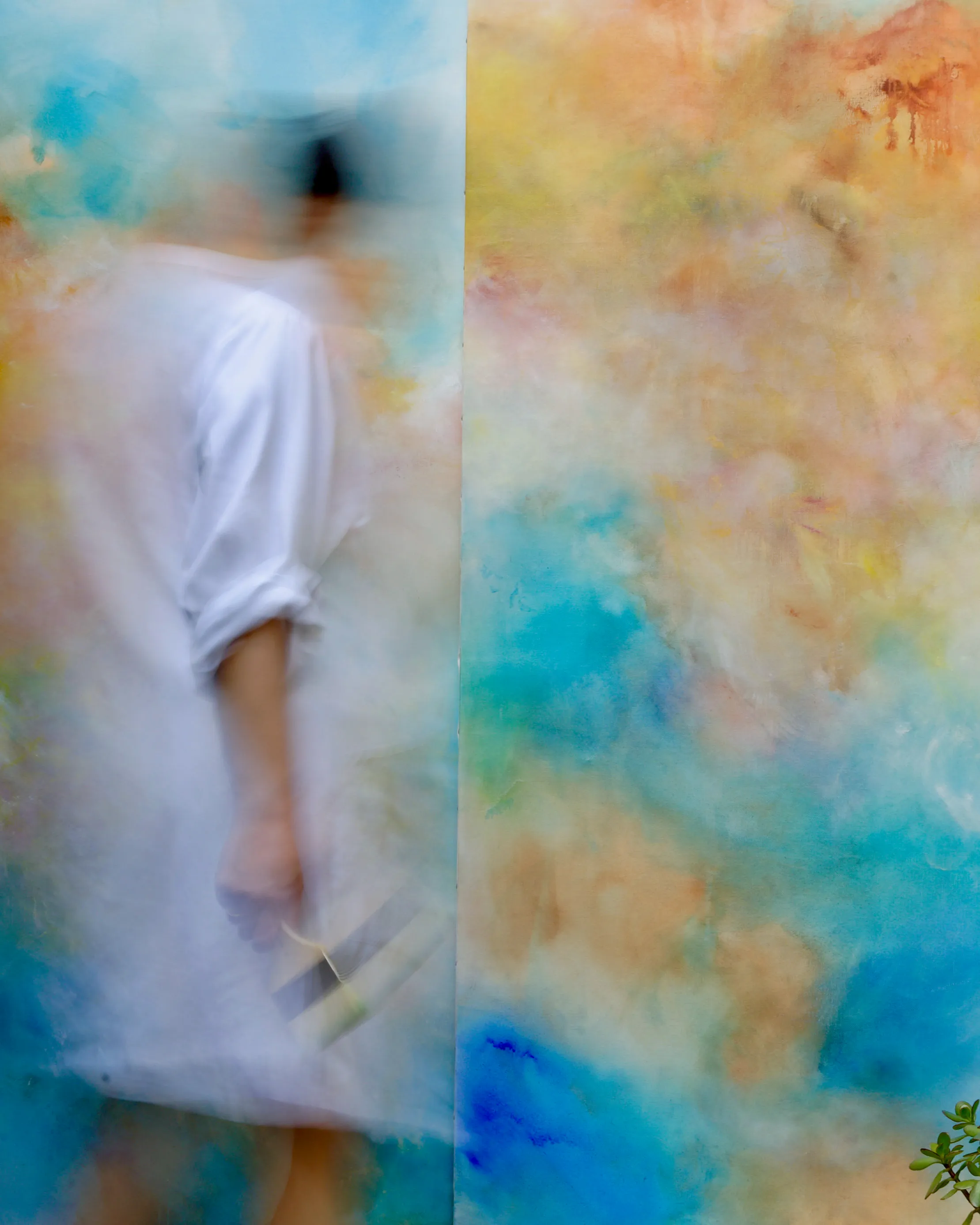 Silhouette floue de l'artiste peintre Mai Khanh Pham To, en blouse de peintre blanche, manches retroussées, de trois-quart dos, un pinceau à la main. Elle est devant une très grande toile colorée dans les tons d'orange, bleus et rosés.