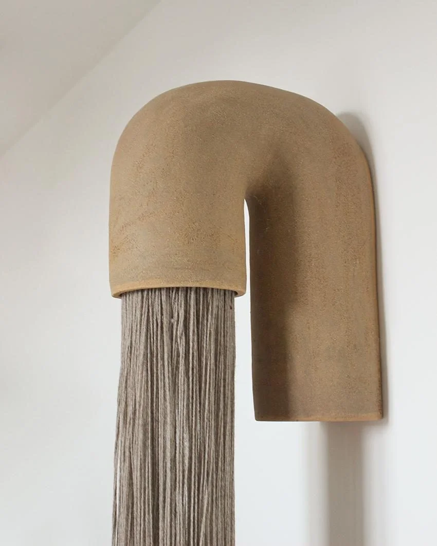 Applique Fontaine #1 d'Elisa Uberti en grès gingerbread, forme en arche d'où tombe une frange de fils beiges, fixée au mur.