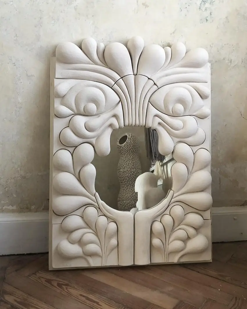 Miroir Oria #1 d'Elisa Uberti en grès blanc, cadre sculpté en forme de visage stylisé aux motifs de plumes et de larmes, adossé à un mur patiné.