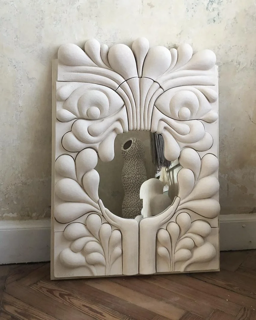 Miroir Oria #1 d'Elisa Uberti en grès blanc, cadre sculpté en forme de visage stylisé aux motifs de plumes et de larmes, adossé à un mur patiné.